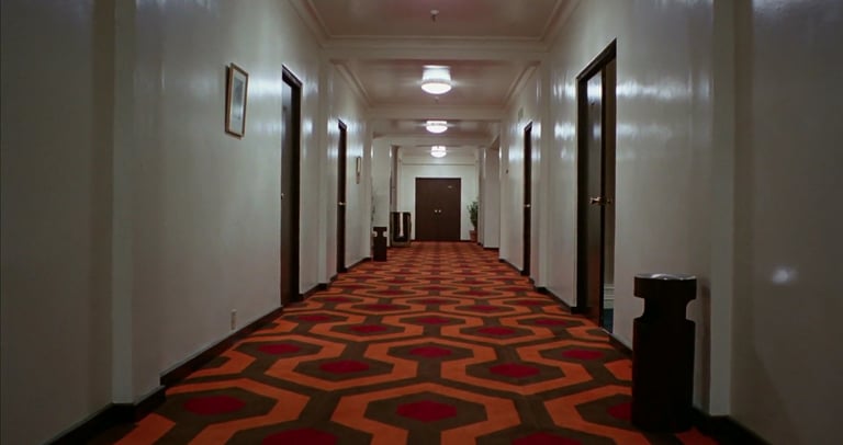 Symmetrical hallway with white walls and a geometric red-orange carpet pattern from The Shining.